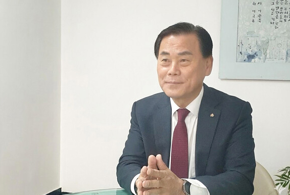 정장을 입은 한 남성이 책상 앞에서 손을 모은 채 인터뷰하듯 앉아 있는 모습으로, 새마을금고 관련 선거와 후보자 인터뷰 장면을 연상시키는 이미지.